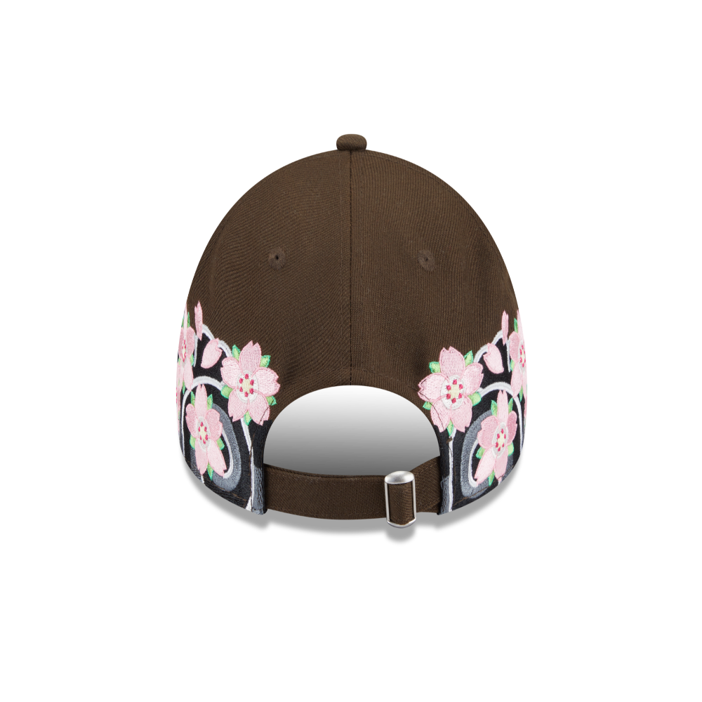 A brown baseball cap shown from the back, featuring a detailed graphic print of pink cherry blossoms and black bicycles along the lower edge, with a silver metal slider on the back strap.