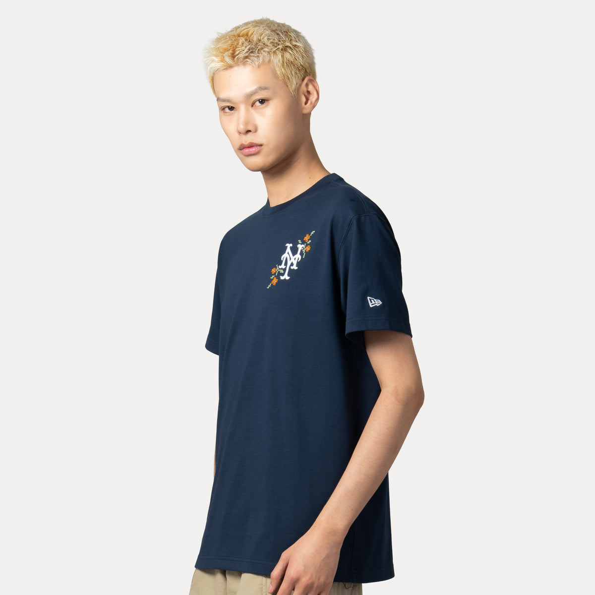 A man wearing a navy blue New Era t-shirt with a white and orange New York Mets logo on the chest, photographed against a light gray studio background.
