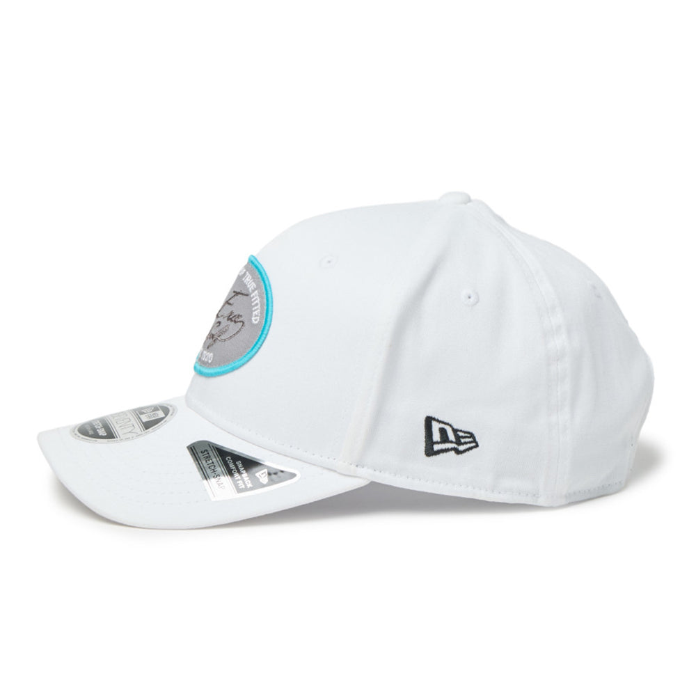 new era script oval patch white golf 9seventy stretch snapback cap with a curved brim, featuring a black embroidered New Era logo on the side and a circular gray and turquoise patch on the front.