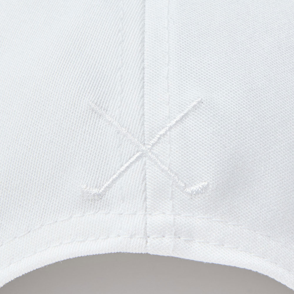 Close-up of a new era script oval patch white golf 9fifty stretch snapback cap showing fine woven texture, a central seam, and a subtle embroidered crossed-stick logo near the center.