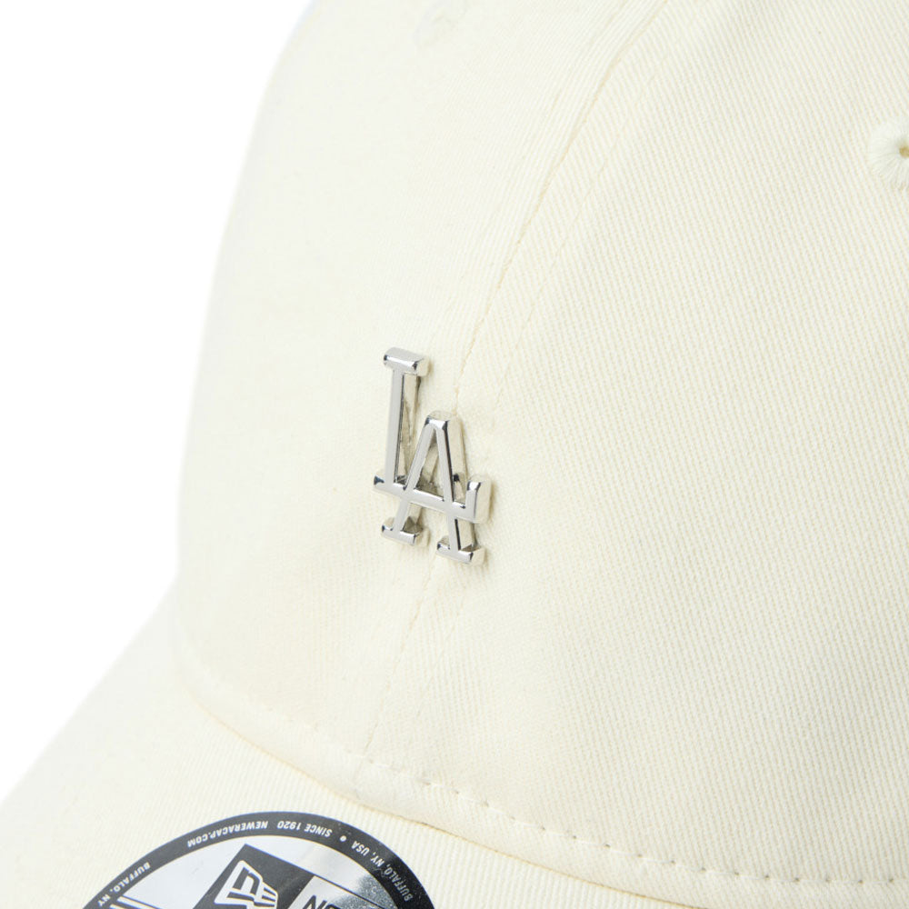 Close-up of a cream-colored baseball new era script mini metal logo cream 9twenty adjustable cap featuring a silver metallic interlocking LA logo on the front and a New Era sticker on the brim.