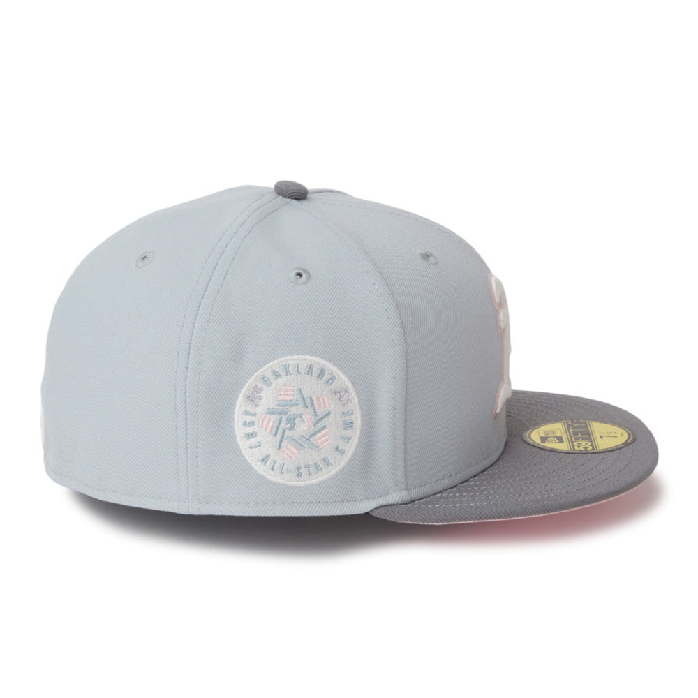A light gray fitted baseball cap with a dark gray visor, featuring a circular embroidered patch on the front and a gold size sticker on the side, set against a plain white background.