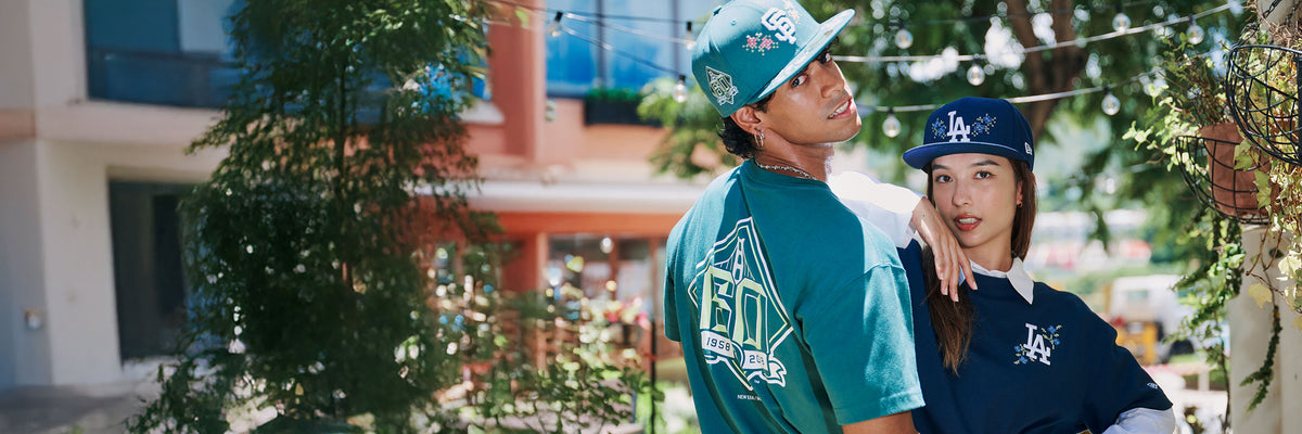 Two people wearing baseball caps in an outdoor setting with trees and buildings in the background.